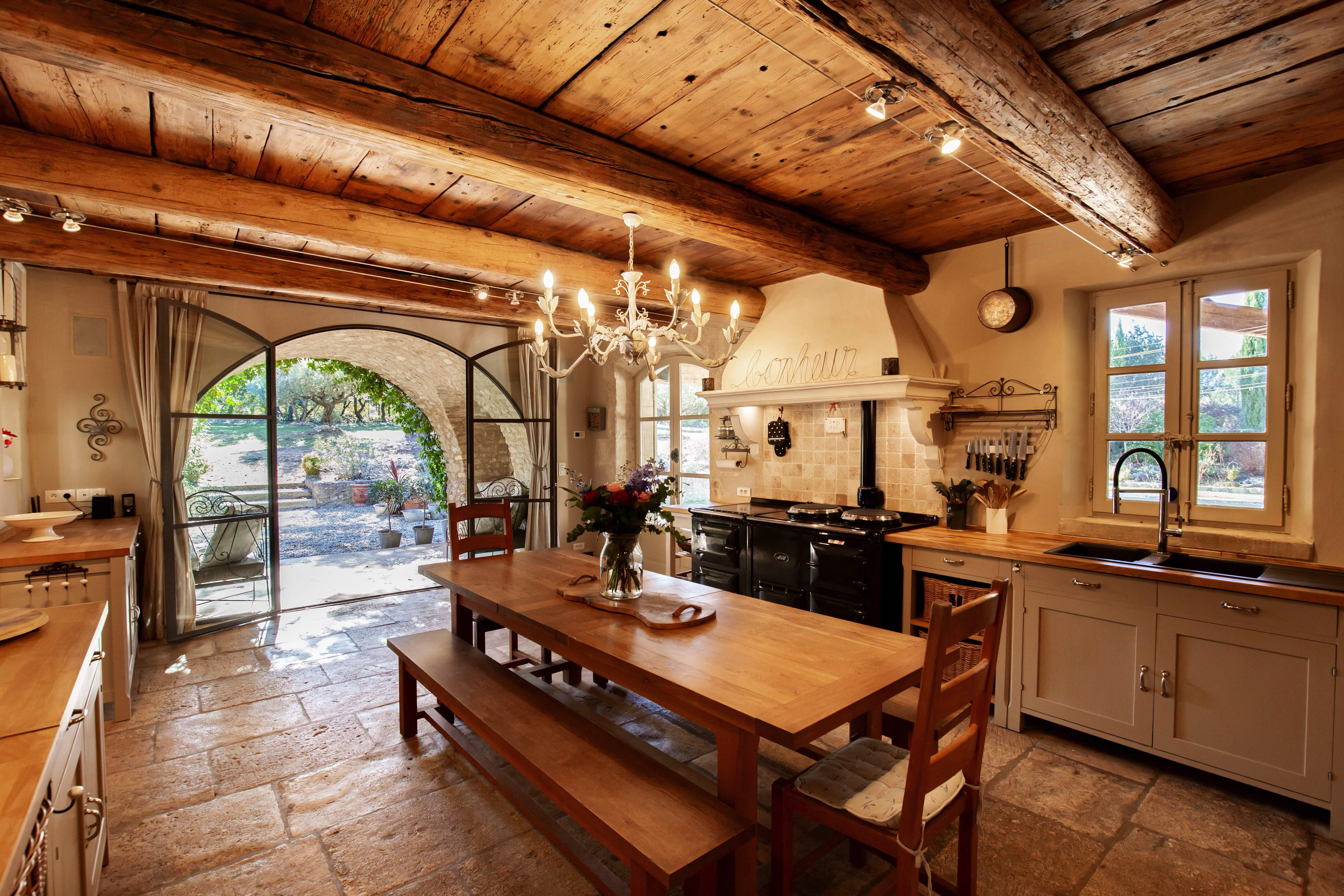 A rustic kitchen with a wooden ceiling and beams, featuring a large dining table, a chandelier, and modern appliances. French doors open to a courtyard.